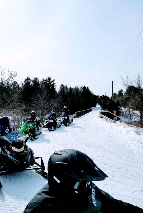 A group of snowmobiles parked on a snowy trail, with trees and a bridge in the background under a clear sky.