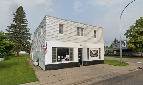A two-story white building with large windows, an American flag, and a grassy area in front.