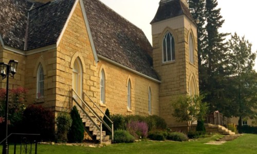 A stone church with a steep roof and tall windows, surrounded by greenery and flowers on a sunny day.