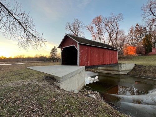 A red covered bridge over a calm stream, surrounded by trees and a sunset sky.