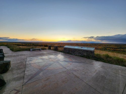 A scenic view at sunset from a lookout point, featuring a compass design on the ground and distant mountains.