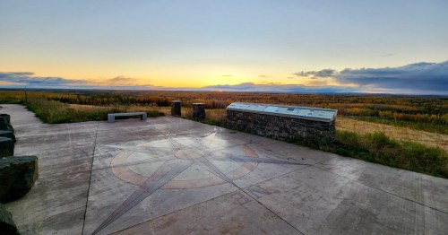 Scenic overlook with a compass rose design on the ground, showcasing a sunset over a landscape of trees and hills.
