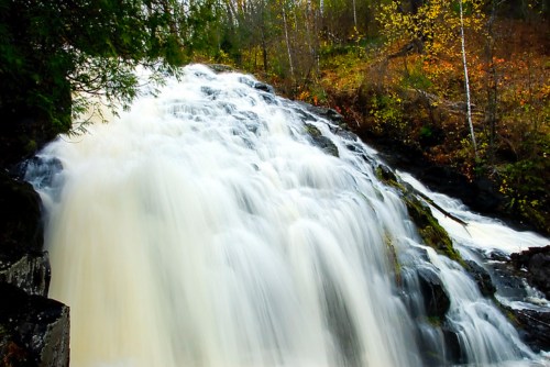 A cascading waterfall flows over rocky terrain, surrounded by lush greenery and autumn-colored trees.