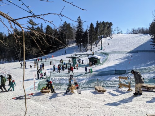 A busy ski slope with people skiing, snowboarding, and enjoying the winter day under a clear blue sky.