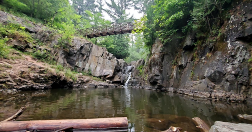 A serene natural scene featuring a small waterfall, rocky cliffs, and a wooden bridge surrounded by lush greenery.