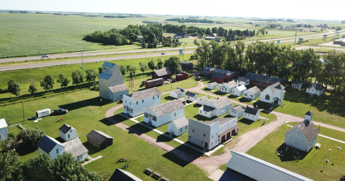 Aerial view of a rural village with various buildings and green fields, surrounded by a highway and trees.