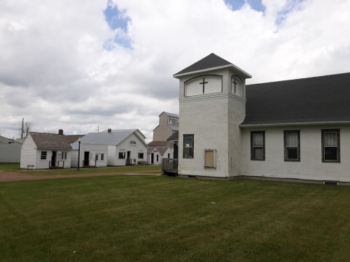 A white church with a tower and cross, surrounded by grassy areas and other buildings under a cloudy sky.