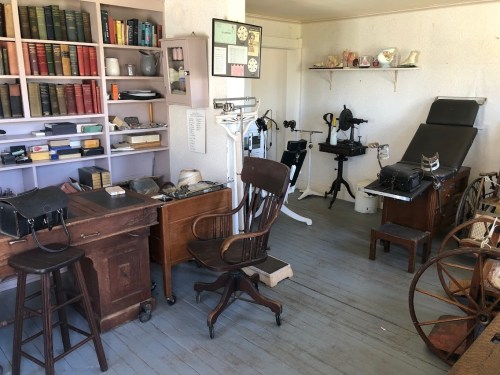 A vintage room with wooden furniture, bookshelves, medical equipment, and a sewing machine, evoking a historical atmosphere.