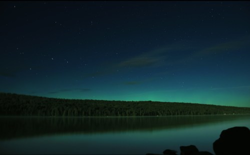 A serene night scene over a calm lake, with a starry sky and a greenish glow reflecting on the water.