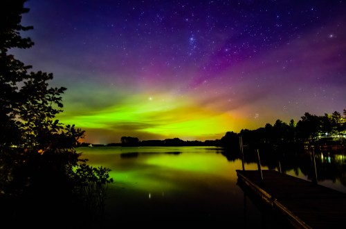 A serene lake reflects vibrant auroras and a starry sky, framed by trees and a wooden dock.