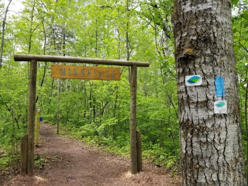 Wooden archway with "Minnesota" sign at the entrance of a forest trail, surrounded by lush green trees.