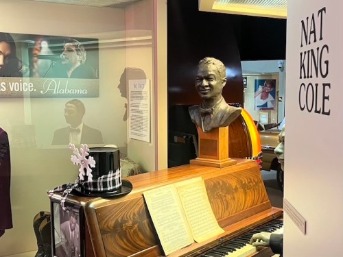 A bust of Nat King Cole sits atop a piano, with a top hat and sheet music nearby, in a museum display.