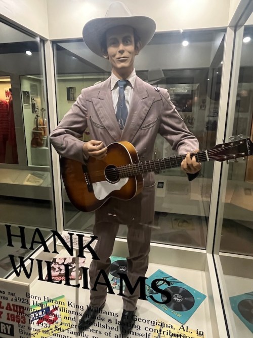 A life-sized statue of Hank Williams in a suit, holding a guitar, displayed in a glass case with memorabilia.