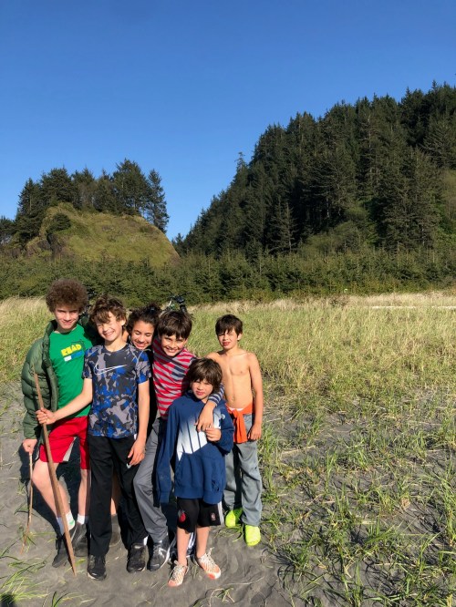 A group of six children stands together on a beach, surrounded by tall grass and trees under a clear blue sky.