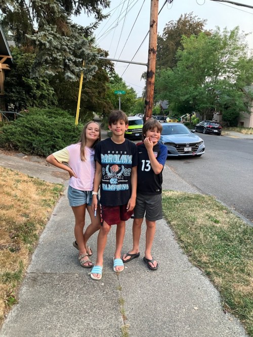 Three kids stand on a sidewalk, smiling and posing for the camera, with trees and cars in the background.
