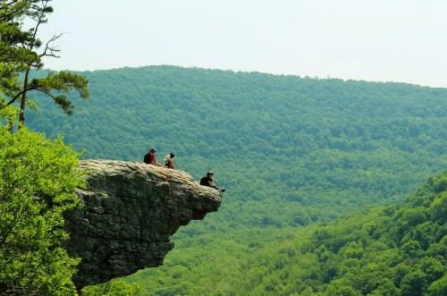 Four people sit on a rocky ledge overlooking a lush green forested landscape under a clear sky.