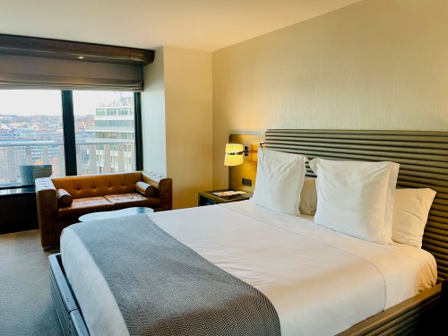 A modern hotel room featuring a king-sized bed, a cozy seating area, and large windows with city views.