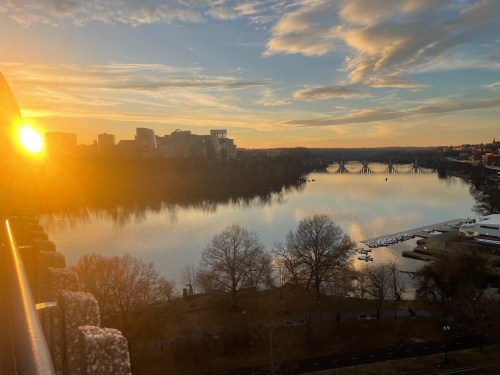 Sunset over a river, reflecting golden light, with trees and buildings in the background. Peaceful and scenic view.