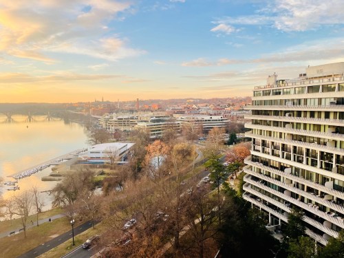 A scenic view of a riverfront city at sunset, featuring modern buildings and trees along the water's edge.