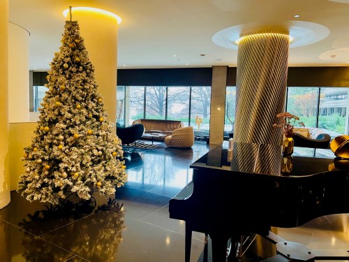 A decorated Christmas tree stands next to a black piano in a modern, stylish lobby with large windows.