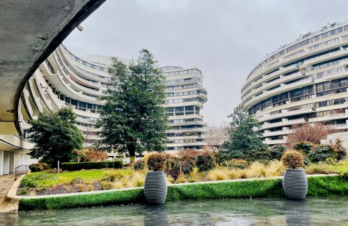 Modern curved buildings surrounded by greenery and water, under a cloudy sky.
