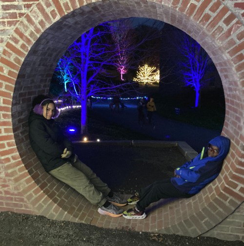 Two children sit in a circular brick arch, surrounded by colorful lights and trees in a festive outdoor setting.