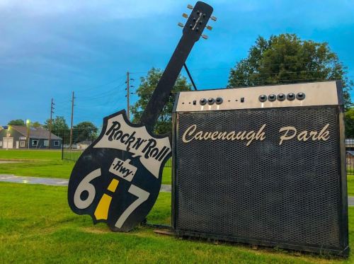 Sign featuring a large guitar and amplifier, promoting "Rock 'n' Roll Hwy 6 & 7" at Cavenaugh Park. Green grass and trees in background.