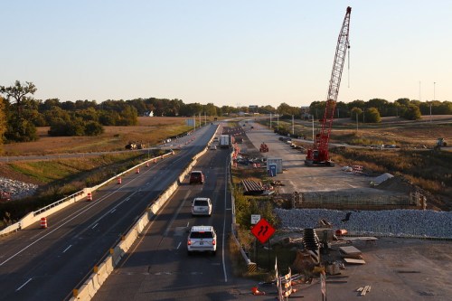 Construction zone on a highway with traffic cones, vehicles, and a crane in the background under a clear sky.