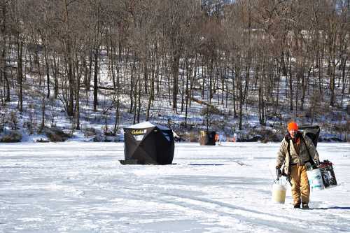 A person in winter gear walks on a frozen lake, carrying supplies, with ice fishing shelters in the background.