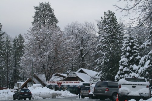 A snowy scene featuring a restaurant sign, surrounded by snow-covered trees and parked cars.
