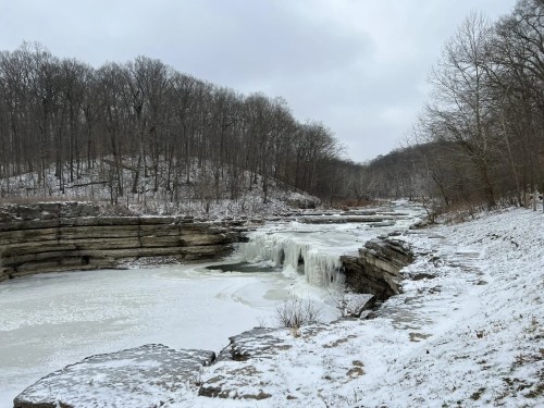 A snowy landscape featuring a frozen waterfall and icy river, surrounded by bare trees and rocky terrain.