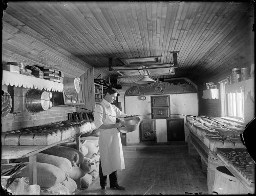 A baker in an old-fashioned bakery, preparing dough with shelves of bread and sacks of flour in the background.