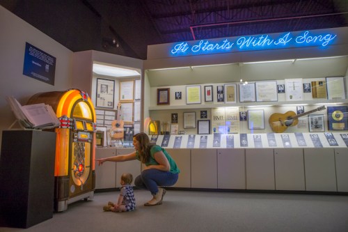 A woman and a toddler interact with a jukebox in a museum, surrounded by music memorabilia and a neon sign.
