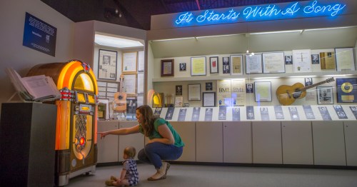 A woman points at a jukebox while a baby sits on the floor, surrounded by music memorabilia in a museum exhibit.