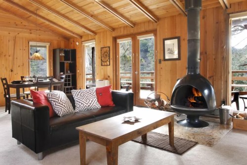 Cozy wooden cabin interior with a fireplace, sofa, dining area, and large windows overlooking nature.