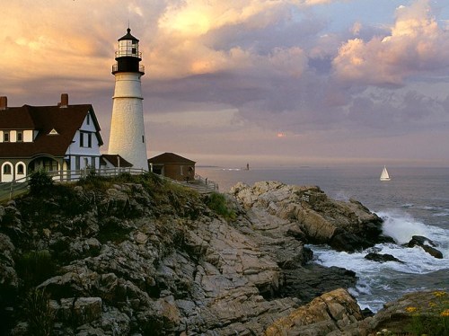 A lighthouse stands on rocky cliffs by the ocean, with a sailboat in the distance and a colorful sunset sky.