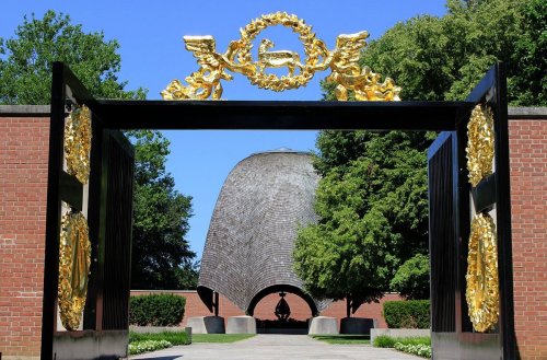 A golden archway frames a modern, curved building surrounded by greenery under a clear blue sky.