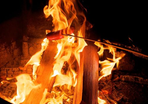 A skewer with a hot dog is held over a roaring campfire, surrounded by bright flames and glowing embers.