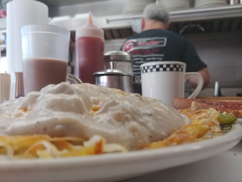 A plate of cheesy breakfast hash with gravy, coffee, and condiments in a diner setting. A cook is visible in the background.
