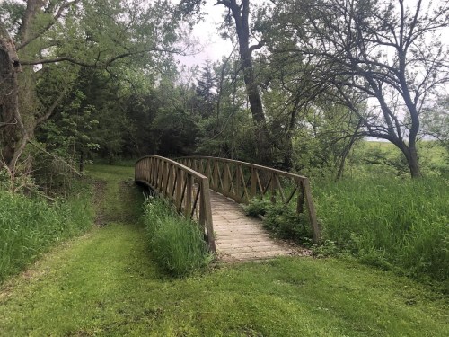A wooden bridge arches over a grassy path, surrounded by lush trees and greenery.