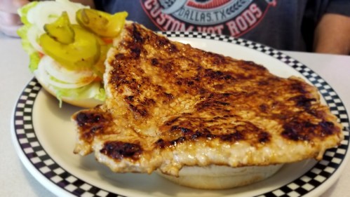 A grilled fish fillet on a bun, served with a side of pickles and a fresh salad.