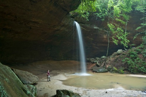 A person stands near a serene waterfall cascading into a small pool, surrounded by lush greenery and rocky cliffs.
