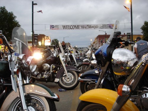 A bustling motorcycle rally with various bikes parked under a "Welcome to Sturgis" banner on a cloudy day.