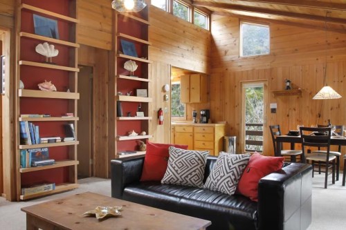 Cozy wooden interior with a black sofa, decorative pillows, bookshelves, and a dining area in a bright, inviting space.