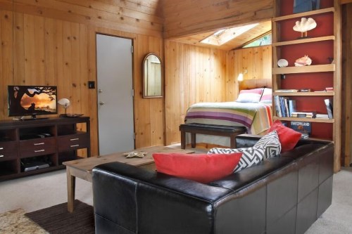 Cozy wooden cabin interior featuring a bed, TV, coffee table, and colorful decor. Natural light from skylight.