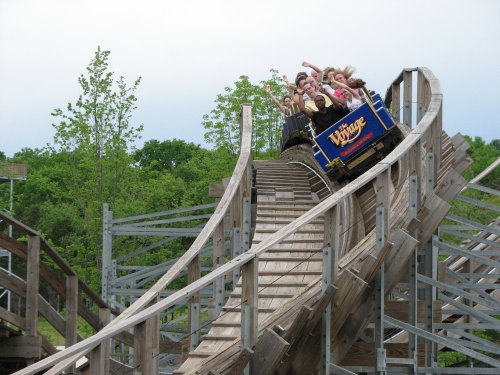 A wooden roller coaster with excited riders going down a steep drop, surrounded by green trees and a cloudy sky.