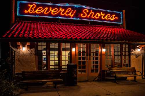 Neon sign reading "Beverly Shores" above a building entrance, with benches and warm lighting at night.