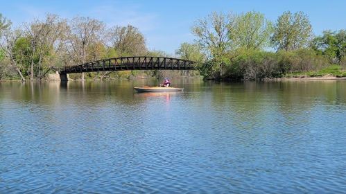 A person kayaking on a calm river with a bridge and trees in the background under a clear blue sky.