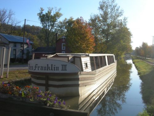 A canal boat named "Ben Franklin III" moored beside a tree-lined path, reflecting in calm water under a clear sky.