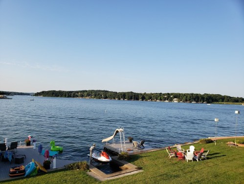 A serene lake view with a dock, colorful floats, a waterslide, and chairs on the shore under a clear blue sky.